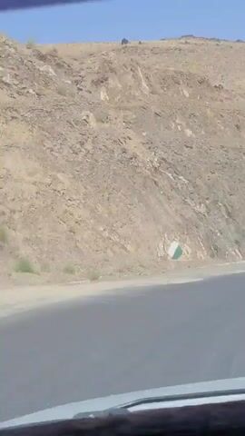 The video shows a car's dashboard and windshield from an interior view. The car is moving along a paved road that curves to the left. To the left of the road, a rocky, barren hillside rises steeply. The ground is dry and covered with sparse, light-colored vegetation. Further up the hillside, there is a white and green road sign. To the right of the road, the landscape consists of more rocky hills and mountains. The sky is clear and bright blue. The terrain appears to be arid and mountainous.