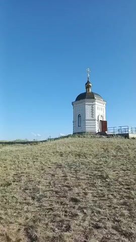 The video shows a panoramic view from a hilltop overlooking a vast, rolling landscape under a clear blue sky. The foreground is dominated by dry, yellowed grass and some sparse green vegetation. To the left, a solitary cross stands on a rocky outcrop. Further in the distance, the terrain unfolds into gently sloping hills and valleys, with scattered trees and what appears to be a small settlement in one of the valleys. The camera pans slowly to the right, revealing more of the undulating countryside. The lighting suggests it is daytime, likely late morning or afternoon, with the sun casting soft shadows. There are no people visible in the frame. The overall impression is one of a remote and serene natural environment.
