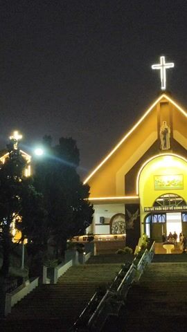 At night, a brightly illuminated church stands out against the dark sky. The church has a large, illuminated cross at its peak and is decorated with warm, glowing lights that outline its architecture. A statue is visible above the entrance, and people are seen entering the church. A long set of steps leads up to the entrance, with a metal railing on the right side. Trees are visible to the left of the church.