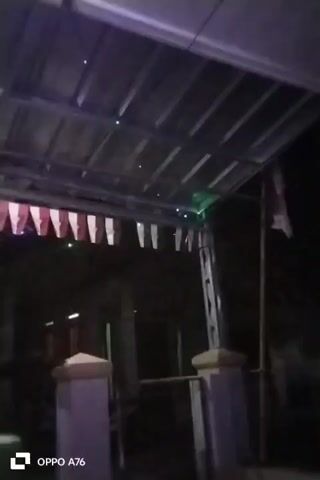 The video shows a night scene outdoors. There is a covered walkway with a corrugated metal roof. String lights, some green and some blue, are hung under the roof. A string of red and white pennant flags hangs along the edge of the walkway. To the right, a flagpole with a red and white flag is visible. The lower part of the frame shows two square posts, one in the foreground, painted purple and cream, and another further back. The image is dark and grainy, suggesting it was filmed in low light conditions. The text "OPPO A76" is visible in the bottom left corner, indicating the device used to record the video.