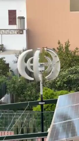 The video shows a small, white, vertical axis wind turbine spinning on a pole, next to a solar panel. The turbine is cylindrical with curved blades around its circumference. The pole is black with silver colored segments. The solar panel is bluish gray, with a grid pattern visible. The background includes a beige building with a brown window, a white building with a dark gray trash can on a balcony, green trees, and a green wire fence.  The turbine is spinning at a moderate speed.