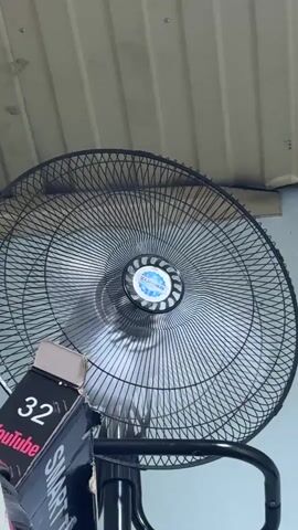 The video shows a close-up of a black oscillating fan with a blue and white logo in the center. The fan is in motion, with its blades blurred from rotation. A cardboard box with "32" and "YouTube" printed on it is visible in the lower left corner of the frame, partially obscuring the fan's stand. The background appears to be a ceiling with some corrugated material and a wall. The lighting suggests it could be daytime, indoors. There are no people visible in the frame. The overall scene is simple, focusing on the fan and the box.