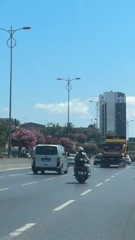 The video shows a view from a vehicle driving on a multi-lane road under a clear blue sky with a few scattered clouds. In the foreground, the right side of a white van is visible. The road has white lane markings. Several vehicles are on the road, including a white minivan, a dump truck with an orange bed, and two motorcycles, each with one or two riders wearing helmets. In the background, there are trees with pink flowers, other buildings, and a tall, modern white building with dark accents. Streetlights are also visible. The traffic appears to be moving.