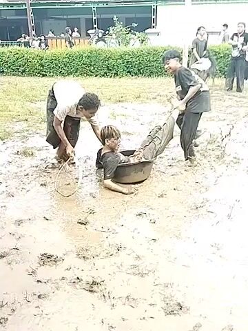 Two people drag a man through mud with a hula hoop. The men are wearing long skirts, one black and the other brown. The man being dragged wears a black shirt and pants. The mud is brown and thick. There are other people in the background, some with drums, watching the spectacle. The people are on a grass field. In the background is a building.