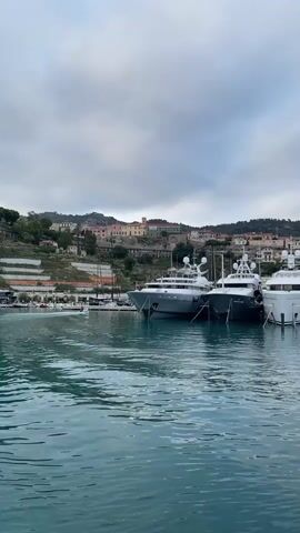 A video clip opens with a view of a harbor with several large yachts docked. The water in the foreground is a deep teal color with visible ripples. In the background, a hillside is covered with buildings and trees, suggesting a coastal town. The sky is overcast with clouds. As the video progresses, the camera appears to move slightly, offering a subtly shifting perspective of the docked yachts and the town. There are no visible people in the immediate foreground, though small boats and figures might be discernible at a distance on the docks or in the water, it is difficult to ascertain details with certainty due to the resolution. The overall impression is a serene, albeit cloudy, day at a well-appointed marina.