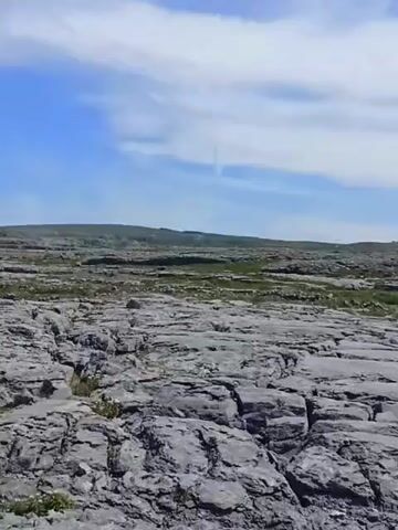 The video shows a barren, rocky landscape under a partly cloudy sky. The foreground is dominated by large, grey, flat rocks with visible cracks and fissures. In the middle ground, the rocky terrain continues, with sparse patches of green grass and low vegetation interspersed. Further in the distance, a low, undulating horizon is visible, also appearing rocky and sparsely vegetated. A road can be seen cutting across the middle ground, and two dark-colored cars are driving on it. The overall impression is one of a rugged, undeveloped, and somewhat desolate natural environment.