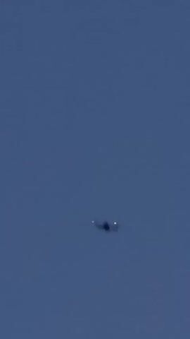 A drone is visible flying in a dark blue sky. The drone is small and appears to be a quadcopter. In some frames, the drone has green lights visible. Below the drone, there are what appear to be illuminated structures, possibly part of a stage or art installation. The structures are covered in reflective material that sparkles. The sky is clear with no clouds.