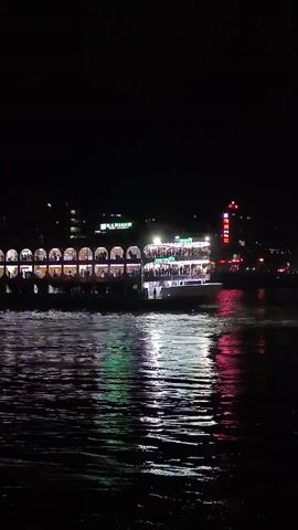 The video shows a large riverboat, illuminated at night, moving on a body of water. The boat has multiple decks visible with arched windows on the lower levels, where silhouettes of people can be seen. Above, the boat is decorated with lights. The reflections of the boat's lights shimmer on the water's surface. In the background, city lights can be seen.