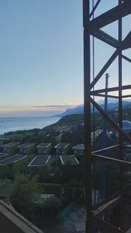 The video shows a landscape with a view of the sea and a series of small, green-roofed buildings, likely villas or houses. The scene is shot from an elevated position, partially obstructed by a metal framework. The sky is a light blue with hints of sunrise, while the sea is calm. Trees and other vegetation surround the buildings. A faint coastline can be seen in the distance, partially obscured by clouds. The overall impression is one of a coastal resort or residential area.