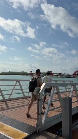 The video begins with a view of two people walking on a wooden pier. The person in front is wearing a backpack and shorts, and the person behind is wearing a light-colored shirt and pants, possibly an employee with a lanyard. In the background, several red and white seaplanes are docked on the water. The sky is blue with scattered white clouds. As the video progresses, the perspective shifts to show more of the seaplanes and the clear turquoise water. A yellow seaplane is prominent in the foreground, with other seaplanes, mostly red and white, visible behind it. The pier has a white metal railing. The video ends with a clear view of the seaplanes against the bright sky.