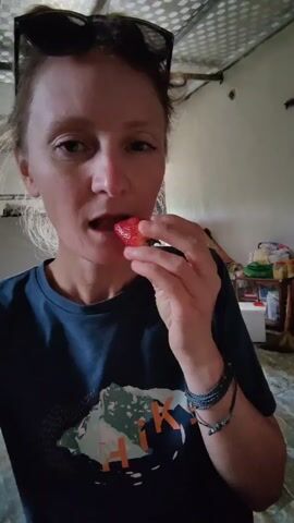 The video features a woman taking a bite out of a strawberry and then showing the bitten strawberry to the camera. She is wearing a dark blue t-shirt with a graphic design, has sunglasses on her head, and several bracelets on her left wrist. Her hair is pulled back, and she is looking directly at the camera. The background is somewhat cluttered, with glimpses of a white cabinet or storage unit, various containers, and a broom visible. The ceiling has a textured, possibly corrugated, appearance. The lighting is somewhat dim.