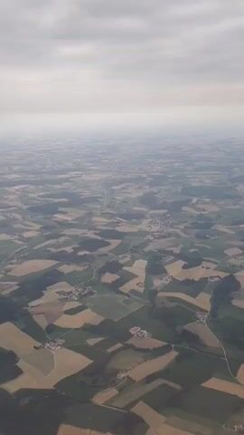 This is an aerial shot of a rural landscape with patchwork fields and scattered houses. The sky is overcast with clouds. The fields are a mix of green and harvested golden crops, suggesting an agricultural region. There are small clusters of buildings, indicative of villages or hamlets, interspersed within the fields. Roads and small tracks can be seen winding through the terrain, connecting the settlements and fields. The overall impression is one of a vast, cultivated expanse seen from high above.