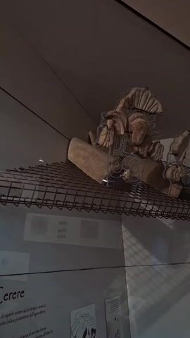 The video pans across an exhibit in a museum. The primary objects are ancient terracotta architectural fragments, possibly roof tiles or decorations, displayed on a wire shelf behind a glass barrier. These fragments feature sculpted faces and decorative motifs, some with remnants of paint. In the background, there are information panels with text and illustrations. The lighting is dim, characteristic of a museum setting. The perspective suggests the camera is moving horizontally from left to right, capturing different pieces of the artifact.