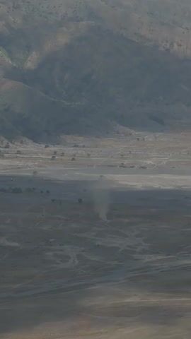 A wide shot captures a vast, dusty valley framed by barren, mountainous terrain. The landscape is characterized by arid, sandy plains with sparse vegetation and winding dirt tracks. In the center of the frame, a dust devil, a swirling vortex of air and dust, is actively forming and spinning. The surrounding hills are covered in a muted, earthy palette of browns and grays, with shadows cast by the sun highlighting the rugged topography. The overall atmosphere is one of dryness and desolation.