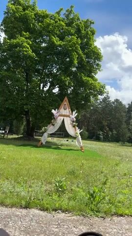 An outdoor static shot captures a wooden A-frame arch draped with white fabric and adorned with pink and white flowers and feathery plumes. The arch stands on a patch of artificial green turf in a grassy field. A large, green-leafed tree is visible directly behind the arch, and a dense treeline extends across the distant background. The sky above is partly cloudy. The immediate foreground shows a rough, dark ground surface. No people or animals are present in the frame.
