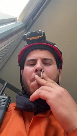A man wearing a red hardhat with a light on top and an orange work suit, is smoking a cigarette and exhaling a cloud of smoke towards the camera. He has a black scarf or neck gaiter pulled up around his neck. He holds a black walkie-talkie in his left hand. The camera is positioned below, looking up at the man. The background shows part of a metal structure or ceiling. He appears to be outdoors or in a semi-enclosed space, with bright daylight filtering in. There is some indistinct audio of speech in the background.