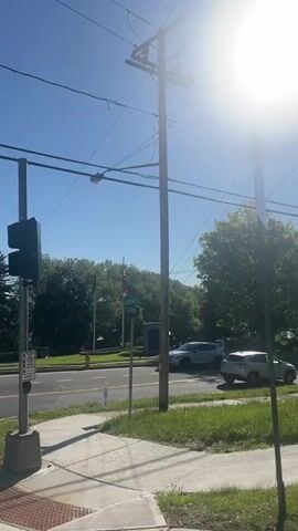 The video shows a sunny street scene. A four-lane road with two lanes in each direction is visible, with cars driving by. There is a sidewalk on the side of the road where the video is taken. Green grass is present between the sidewalk and the road. Power lines and utility poles are visible in the background, partially obscured by the bright sun. Trees are also present, partially obscuring a house in the distance. A street sign is visible, reading "Liberty."