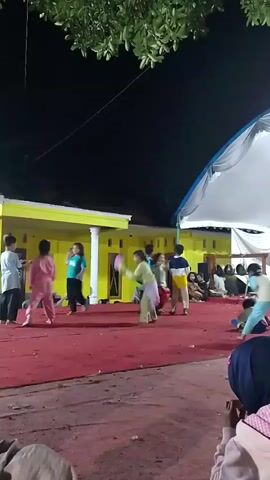 A group of children are dancing on a red carpet stage at night. The stage is in front of a yellow building with white pillars. A white tent is set up to the right of the stage. Several children are actively dancing, some in the foreground and others in the background. Some of the children are wearing brightly colored clothing. There are spectators seated to the right of the stage, also at night.