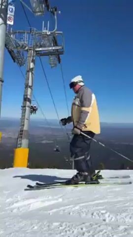 The video shows a ski lift on a sunny day, with skiers ascending the mountain. The sky is clear and blue. In the background is a mountain range and a valley. The ski slope is covered in white snow. A lone skier comes down the slope.