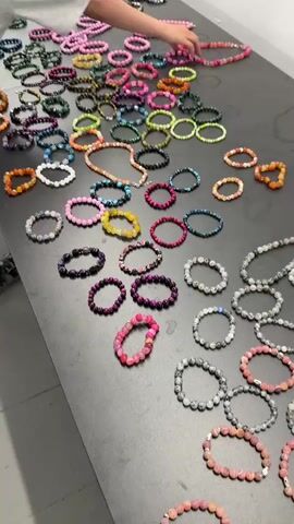 A young person is standing at a long black table on which many colorful beaded bracelets and necklaces are laid out. They are holding up a pink, orange, and blue beaded bracelet and then lay it back down on the table. The person is wearing a light gray t-shirt, gray shorts, and black Crocs. The lighting in the room is bright.