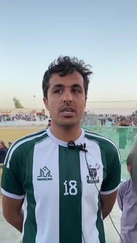 The video shows a close-up, slightly upward-angled shot of a man in a green and white striped soccer jersey with the number 18 on the front. He has a microphone attached to his shirt. He is looking directly at the camera and speaking. In the background, there are spectators in bleachers, a fence, and some trees under a clear sky. The lighting suggests it is daytime, possibly late afternoon or early evening.
