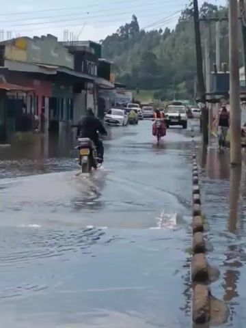 The video shows a flooded street in what appears to be a small town or village. The water level is high enough to submerge parts of the parked vehicles. Two motorcycles are navigating through the flooded street, creating splashes as they move. Other cars and pedestrians are also visible in the background, and there are buildings on either side of the street. The overall atmosphere is one of disruption caused by the flooding.