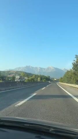The video shows a forward-facing view from a vehicle traveling on a highway. The road is asphalt with white lane markings. On the left side of the road, there is a concrete barrier and then trees and some buildings. On the right side of the road, there is a metal guardrail and trees. In the distance, a mountain range with snow-capped peaks is visible against a clear blue sky. The sun is shining, and the lighting suggests it is daytime. The scene is tranquil and scenic, depicting travel through a mountainous region.