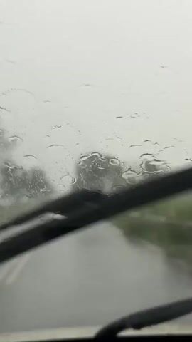 The video shows a view from inside a vehicle, looking out through a wet windshield. Raindrops are visible on the glass, distorting the view of the road ahead. The road is wet and appears to be paved, with a white line marking the center. There are trees and foliage on either side of the road, which are blurred due to the rain and motion. In the distance, the headlights of another vehicle are visible, appearing as blurred yellow lights. The sky is overcast and grey. The windshield wipers are not in motion during the clip. The overall impression is of driving in rainy weather.