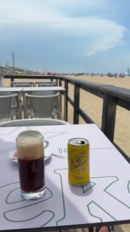 The video begins with a close-up of a table at an outdoor cafe. On the table, there is a glass with a dark beverage topped with foam, a small white saucer with a sugar packet, and an open can of Schweppes Tonic Water. The table has a white surface with a green pattern. In the background, rows of empty white tables and chairs are visible, suggesting a dining area. Beyond the cafe, a sandy beach stretches out with people scattered across it, some sitting under colorful umbrellas. The ocean is visible in the distance under a bright, partly cloudy sky. The camera then pans slightly to the right, offering a wider view of the beach scene. More people are seen on the beach, along with beach umbrellas and tents. The video continues to pan and zoom out, showing more of the expansive beach, the ocean, and the sky. The focus shifts to the beachgoers and the overall atmosphere of a sunny day at the seaside. Towards the end, the video offers a panoramic view of the beach, highlighting the presence of various beach structures, people enjoying the sun, and the vast expanse of sand and sea under a clear blue sky.