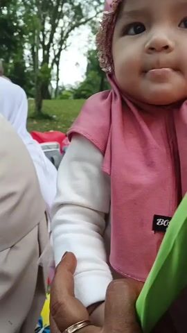 This is a close-up video of a baby wearing a pink hijab with lace trim. The baby has dark eyes and is looking towards the camera. A hand is visible holding the baby's arm. In the background, a person in a white hijab and another person in a black hijab can be seen. There are also some objects that appear to be bottles and a picnic blanket scattered on the grass in the background, suggesting an outdoor setting. The baby's facial expression changes slightly throughout the short clip, showing a pout.