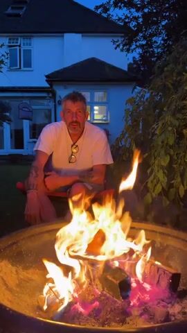 A man sits in a red chair in a garden at dusk, near a fire burning brightly in a circular metal fire pit. The fire pit contains multiple pieces of wood that are engulfed in flames. The man wears a white t-shirt, light-colored shorts, and has glasses hanging around his neck. Tattoos are visible on his arms. A two-story house with white walls and a dark roof stands in the background, with a light illuminated in one of the windows. The garden also contains grass and a tree with long branches.