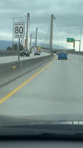 The video shows a first-person perspective from inside a car driving on a multi-lane highway on a cloudy day.  The highway has a concrete median barrier separating the lanes.  A yellow stripe marks the edge of the lane nearest the median.  Other cars and trucks are visible driving in the same direction, with some traffic congestion ahead.  The road is part of a long bridge supported by several tall cable-stayed towers.  A green highway sign is visible in the upper right of the frame.