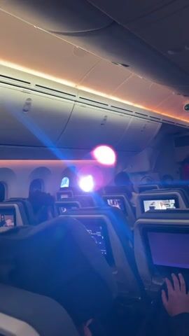 The video shows the interior of an airplane cabin, likely during a flight. Several rows of seats are visible, each equipped with a personal entertainment screen. Passengers are seated in some of the seats, while others appear to be unoccupied. The cabin lighting is a mix of blue and orange hues, creating a unique ambient light. A strong glare or lens flare obscures part of the view. The overhead compartments are closed, and the general atmosphere suggests a comfortable yet somewhat dimmed setting.