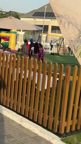The video shows a backyard scene with a brown wooden picket fence in the foreground. Behind the fence, there is a green lawn with several people gathered. To the left, there is a colorful inflatable bouncy castle. Some people are standing and talking near tables covered with white cloths, while others, including children, are playing on a seesaw and other playground equipment. A person dressed in a Mario costume is visible, and a person is holding a child in front of the fence. Several people are holding lighting and camera equipment, suggesting a photoshoot or filming session is taking place. The backdrop includes a house with solar panels on the roof and large white umbrellas providing shade. A palm tree is visible on the right side of the frame, partially obstructing the view.