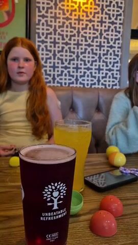 The video shows a family gathered around a table at a restaurant. There are two girls in the video: one with long red hair and a yellow shirt, the other with brown hair and a fuzzy green sweater. There are various drinks on the table, including a dark red pint, a glass of orange soda, and a glass of water with a lemon slice. Additionally, there's a cell phone and some small, colorful plastic toys on the table. The girl in the green sweater is sitting up straight and talking, gesturing with her hands as she speaks. The background features a patterned blue and white wall, a padded booth seat, and a menu attached to the wall. The lighting in the restaurant appears warm, highlighting the wood table.