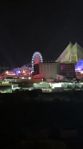 This video shows a nighttime view of a festival or amusement park. In the background, a large Ferris wheel with red lights is rotating. To the right of the Ferris wheel, a large, modern building with a distinctive white, angled roof structure is illuminated with colorful lights, primarily pink and blue. In the foreground and mid-ground, various tents, temporary structures, and buildings are visible, some with lights on. The overall scene is dark, with the illuminated structures and attractions standing out against the night sky. There are no people clearly visible in the frame.