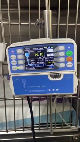 The video shows a small dog in a cage at what appears to be a veterinary clinic or animal hospital. The dog, a Shih Tzu type, is lying on a pink and purple towel. It is wearing a white medical collar and looks towards the camera. Next to the cage is a veterinary infusion pump that is administering fluids. The pump's screen displays settings and progress related to the infusion. The cage is metal and the background consists of stainless steel surfaces.