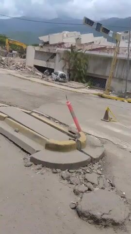 The video shows a street scene with significant destruction. In the background, a large pile of rubble, likely from a collapsed building, dominates the view. A yellow excavator is visible amidst the debris, suggesting ongoing cleanup or demolition efforts. To the right of the rubble, a partially collapsed building stands, with a damaged white car visible in its vicinity.

In the foreground, the street and sidewalk are cracked and damaged, with debris scattered around. A concrete curb with yellow markings is also broken. An orange traffic cone with a white and red striped cap is propped up on the curb. Two women are standing on the sidewalk on the left side of the frame, one wearing a dark top and skirt, the other in a lighter-colored dress. A man wearing an orange vest over a dark uniform and a cap is walking towards the women, possibly a rescue worker or official. The sky is cloudy, and mountains are visible in the distance. The overall impression is one of a disaster zone.