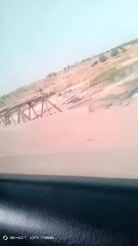 A car driving along a bridge is seen in a video shot from inside the car. The bridge has a white metal railing. Outside the bridge is a dry, desert-like landscape. The camera angle is canted.  A watermark "SHOT ON PG5-" is in the lower left corner.