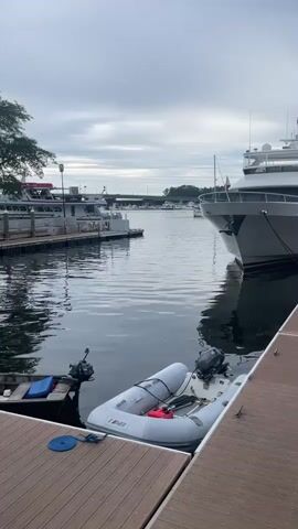 The video shows a marina with several boats docked. The water is calm and reflects the cloudy sky. In the foreground, there is a wooden dock with a small inflatable boat tied to it. In the background, there are larger yachts and ferries docked. Trees line the shore on the left side of the frame. The overall impression is one of a quiet, peaceful morning at a marina.