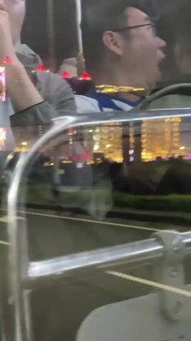 The video shows two men riding on a bus at night. Both are wearing glasses. The man on the right yawns widely at the beginning of the video. The cityscape is visible through the bus window, featuring brightly lit buildings and a tall tower illuminated with colored lights. The video focuses on the scenery visible outside the bus.