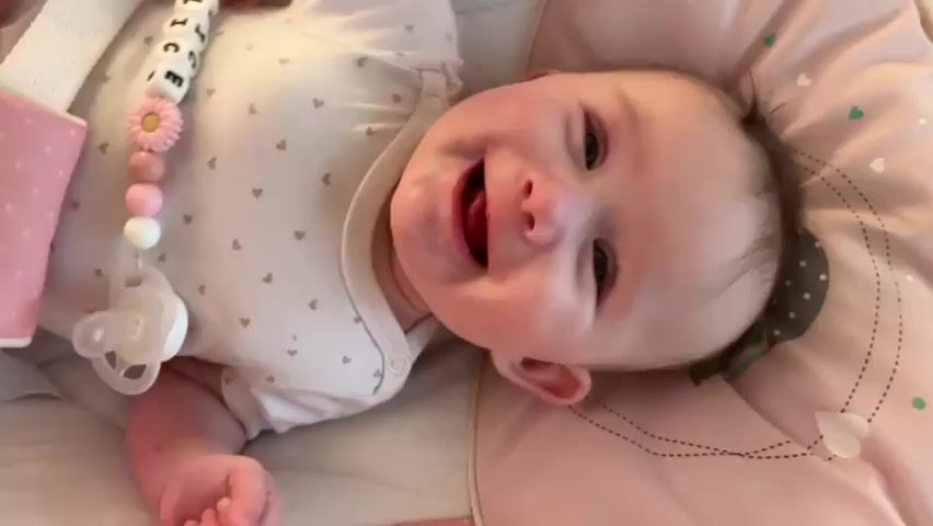 A baby lies on a pink and white cushioned surface. The baby is wearing a light-colored onesie with small heart patterns. A pacifier is attached to a beaded string that also has letters spelling out "ALICE" and a pink flower-shaped bead. The baby is looking up and smiling, with its mouth open. The background is slightly blurred, showing the pink and white cushioned surface and a hint of a white strap on the left. The lighting suggests an indoor environment.