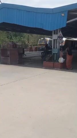 Here is a description of the video:

The video shows a train station with a covered platform. A woman in a red saree stands near a pillar with a sign, then walks to the other side of the platform where there are two trains parked. One train is white and yellow, and the other is a light yellow and blue. A man sits on a bench in the background. The area appears clean and well-maintained.