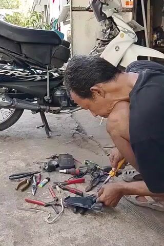 A man with short, dark hair and a cigarette in his mouth kneels on a paved surface, working on a motorcycle engine part with a soldering iron. Numerous tools, including pliers, screwdrivers, and a utility knife, are scattered around him. A black and white motorcycle is parked to his left, and a white motorcycle is visible in the background to his right. The environment appears to be an outdoor workshop or repair area, with a concrete floor and a building structure in the background.