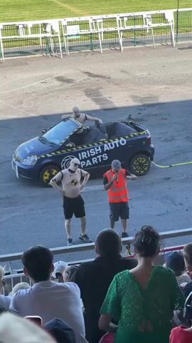 Two male mannequins are positioned in and around a blue convertible car that has been modified with "IRISH AUTO PARTS" branding. One mannequin is seated in the driver's seat and another is standing outside the car to the left. A man in an orange safety vest is speaking into a microphone, facing the standing mannequin. Another man in a green safety vest is looking at a phone in the foreground to the left, and other spectators are seated in the background. The scene appears to be outdoors on an asphalt surface with a grassy area and white barriers in the background, possibly at a race track.