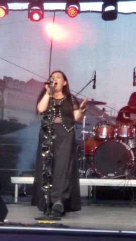 The video is a close-up shot of a female singer performing on a stage at night. She is wearing a black, sleeveless top and a long black skirt with decorative metal accents. The singer has long dark hair and is holding a microphone to her mouth while singing. She is gesturing with her left hand. Behind her, a drum set is visible, along with some stage lighting, including red and white lights that illuminate the stage with a pinkish glow. There is also some haze or smoke on the stage. The background of the stage appears to be a large backdrop with a blurry image of buildings. A part of a guitar neck is visible in the bottom left corner. The video is shot vertically and is slightly shaky.