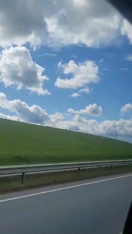 A side view from a vehicle shows a highway with a green grassy hill on the left. The sky is blue with scattered white clouds. A metal guardrail separates the highway from the hill. A blue truck passes by on the highway.