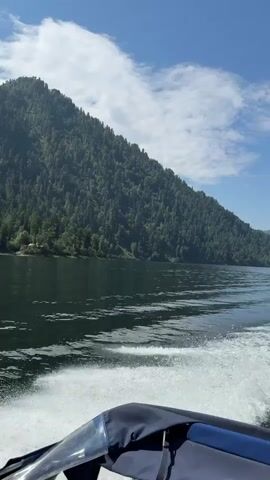 This video shows a view from a moving boat on a body of water. The boat is leaving a white wake behind it. On the left side of the frame, a large, tree-covered mountain slopes down to the water. The sky is blue with some white clouds. In the distance, more mountains can be seen. The camera is pointed slightly downwards and to the right, showing the front of the boat and the water.