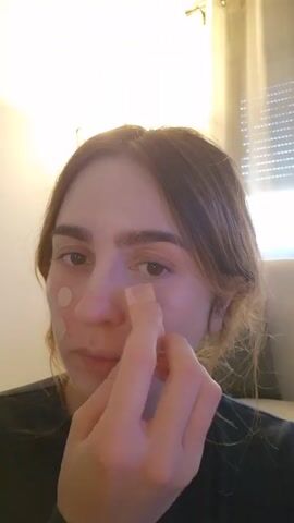 A person with light brown hair is close to the camera, applying a beige cosmetic product to their face. They are wearing a black top. Three circular patches, presumably acne patches, are already on their left cheek. Their right hand is being used to dab the product onto their face. The background shows a white wall with a light fixture and a window with closed blinds.