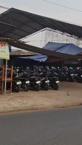 The video shows a parking area filled with motorcycles, most of which are black or dark-colored. There are also a few red motorcycles visible. Some motorcycles have helmets on them. In the background, there are buildings with corrugated metal roofs. A sign on one of the buildings reads "HASAN KOST" and provides an address and phone numbers. Another sign is yellow and displays "PARKIR MOTOR SEPEDA MOTOR Rp. 3.000". A third sign features an image of a golden cigarette and the text "MAGNUM KRETEK Rp. 15.000". The overall scene appears to be an outdoor parking lot, likely associated with a boarding house or kost.